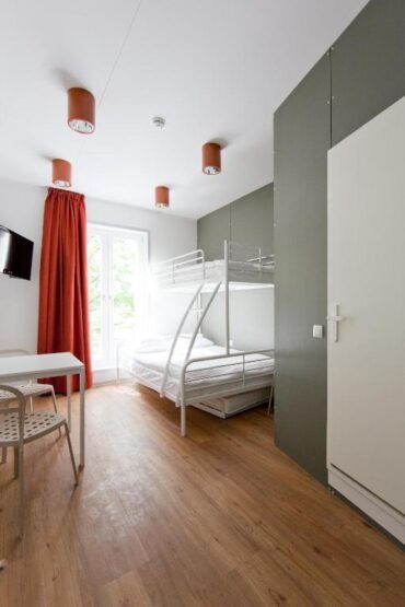 JustResti minimalistlik puitpõrandaga tuba, kus on narivoodi, valge kirjutuslaud, tool, seinale paigaldatud televiisor ja suur punaste kardinatega aken. Ülalt ripuvad kolm pruuni laevalgustit, mis heidavad rohelistele ja valgetele seintele sooja kuma.