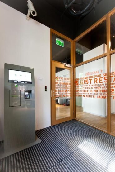 Moodne interjöör, kus vasakul on JustRest iseteeninduskiosk, paremal pool paksu punase tekstiga klaasuks ja lakke paigaldatud turvakaamera. Ruumi täiendavad triibuline vaip ja heledad puidust aktsendid.