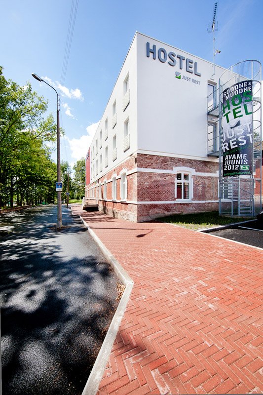 Välisvaade tellistest ja valgest hoonest, millel on sildid HOSTEL, mis asub puudega ääristatud tee kõrval. Metallist keerdtrepp loob huvitava kontakti modernsuse ja looduse vahele. Linnakeskkonnas on selge taevas, mis kutsub avastama.