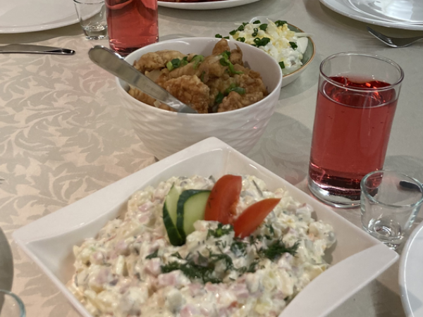 Pikale söögilauale on kaetud valged taldrikud ja klaasides punased joogid. Esiplaanil on kauss salatiga kurkide ja tomatitega ning laua äärde on paigutatud muud toidud, sealhulgas praetud esemed ja köögiviljad.