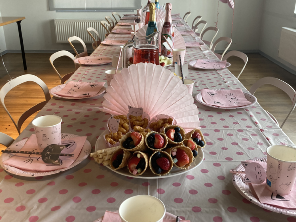 Roosa-teemalise kaunistusega peolaud, mis sisaldab kassi näoga pabertaldrikuid, tasse ja salvrätikuid. Suupisted nagu marjakattega saiakesed on laual. Üleval hõljuvad roosad õhupallid, mis loovad piduliku õhkkonna.