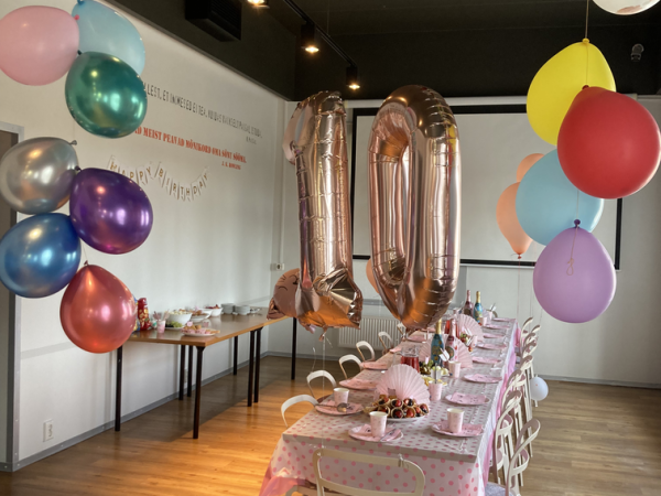 Pidulik peoruum värviliste õhupallide ja suurte roosakuldsete 10 õhupallidega. Pikale lauale on kaetud roosa täpiline laudlina, peomütsid ja taldrikud. Ruumis ripuvad laest eri värvi heledad õhupallid.