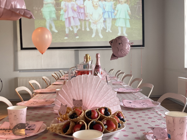 Roosade ja kassiteemaliste kaunistustega pidulik laud peoks. Taldrikutel ja tassidel on kassikujundus. Keskosas on värvilised puuviljaküpsetised. Ülal hõljuvad õhupallid ja suurel ekraanil on väljas värvilistes kleitides lapsed.
