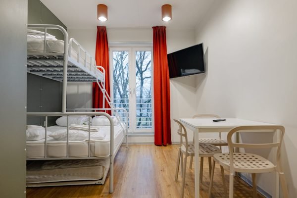 Vaade esikust tuppa, nähtaval on kolmekohaline söögilaud, narivoodi, aken punaste piumendavate kardinatega.